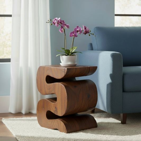 Haussmann Wave solid wood end table in mocha finish, 12 x 14 x 18 inches, sculptural S-curve design with natural knots and fine surface checks in the grain, styled in a living room beside a medium blue sofa with a white ceramic bowl and purple orchid, near a window with white sheer curtains and natural sunlight.